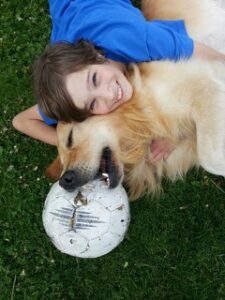 Tochter und Goldenretriver Linus liegen mit einem Fußball auf der Wiese, die Tochter lächelt glücklich